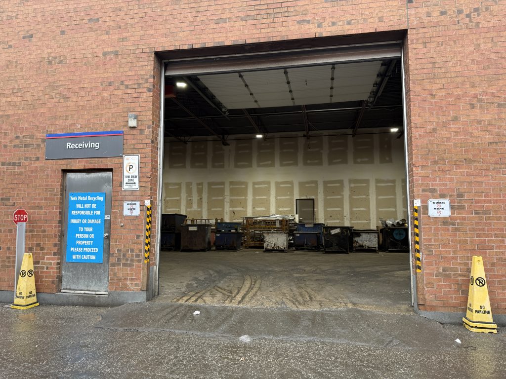 A large open garage door reveals an indoor loading area with stacked materials and several containers. The brick building has “Receiving” signage and yellow caution cones on either side of the entrance.