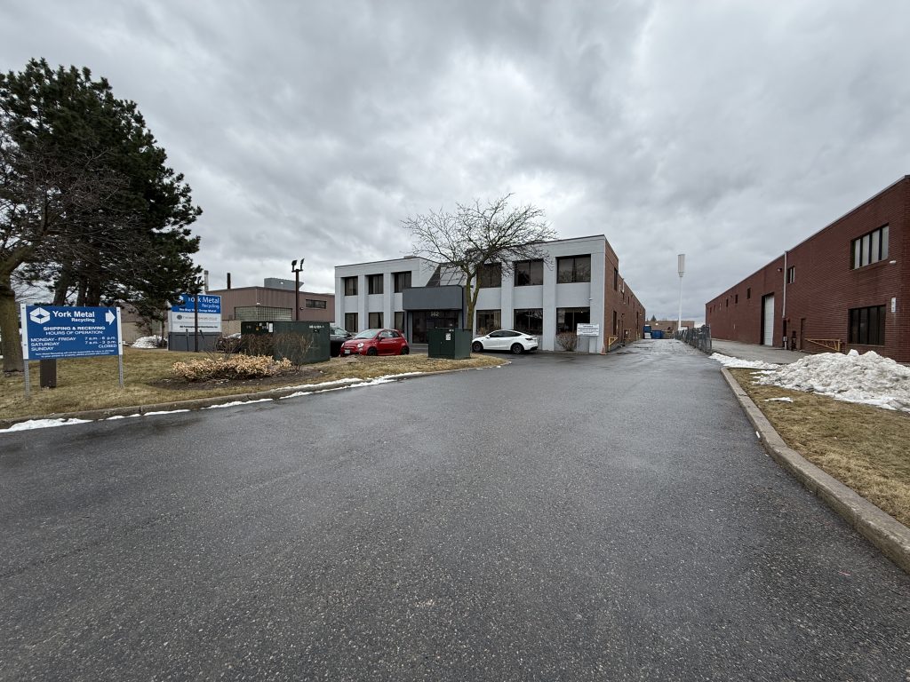 A two-story office building with a tree in front, three parked cars, and a blue sign on the left. The sky is overcast, and the parking lot and sidewalk are wet. Snow patches are visible along the edges.
